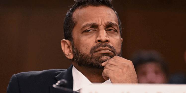 Kash Patel in a suit sits with his hand on his chin, looking skeptical or thoughtful during a formal event. A blurred background and a partial nameplate are visible in the foreground.