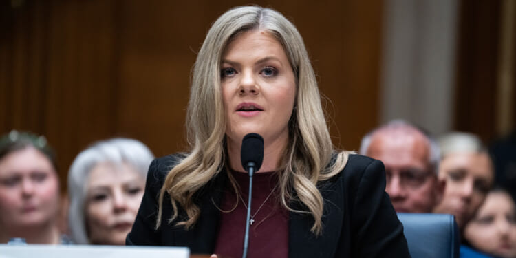 A woman with long blonde hair speaks into a microphone at a formal hearing, wearing a black blazer and maroon top. Several people sit blurred in the background.