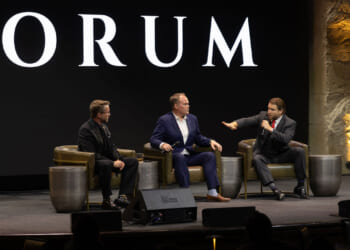 Joe Allen, Matthew Peterson, and Joe Lonsdale in suits sit on stage in armchairs, engaged in a panel discussion. One man gestures while speaking, as the others listen. A large screen behind them displays the word FORUM..