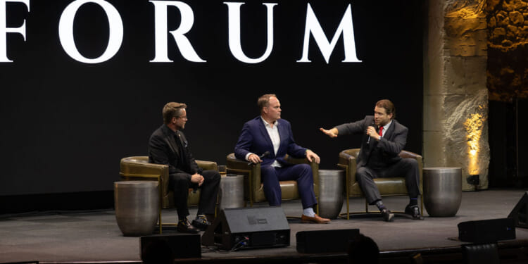 Joe Allen, Matthew Peterson, and Joe Lonsdale in suits sit on stage in armchairs, engaged in a panel discussion. One man gestures while speaking, as the others listen. A large screen behind them displays the word FORUM..