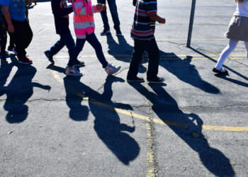 A group of children walk together on a paved surface, casting long shadows in the sunlight. Only their legs and feet are visible, showing various outfits and shoes.