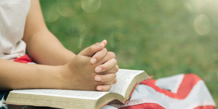 A person sits outdoors with hands clasped in prayer over an open book, draped with an American flag on their lap, with green grass in the background.