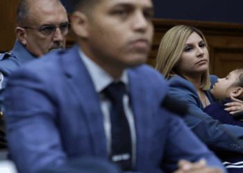 A woman holds a child while seated beside men in suits and uniforms at a formal hearing or meeting, all appear serious and attentive.