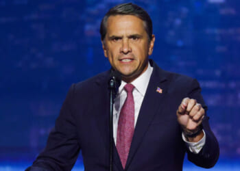 Todd Blanche in a dark suit and red tie speaks passionately at a microphone, gesturing with his right hand, against a blurred blue background.