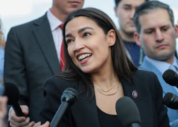 The American Federation of Government Employees, or AFGE, endorsed Rep. Alexandria Ocasio-Cortez and many other Democrats. Pictured: AOC, a woman in a black blazer speaks to reporters, surrounded by microphones and people holding recording devices at an outdoor press event.