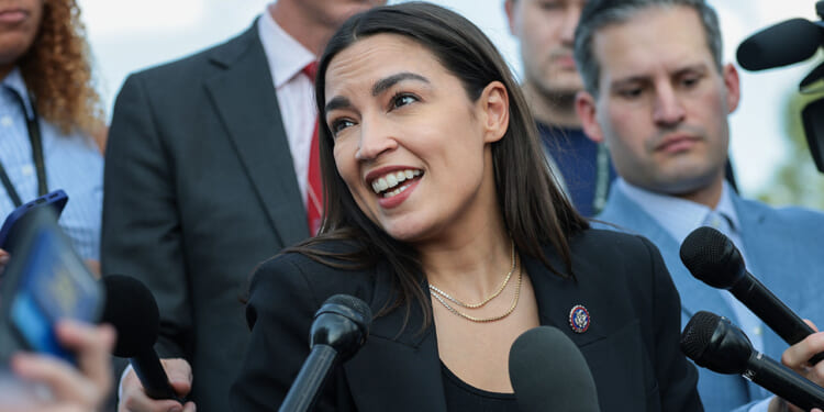 The American Federation of Government Employees, or AFGE, endorsed Rep. Alexandria Ocasio-Cortez and many other Democrats. Pictured: AOC, a woman in a black blazer speaks to reporters, surrounded by microphones and people holding recording devices at an outdoor press event.