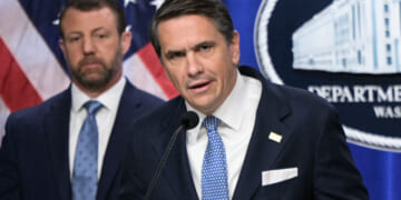 Todd Blanche and Markwayne Mullin in suits stand in front of U.S. flags and a sign that reads Department. The man in front speaks into a microphone, while the other looks on in the background.