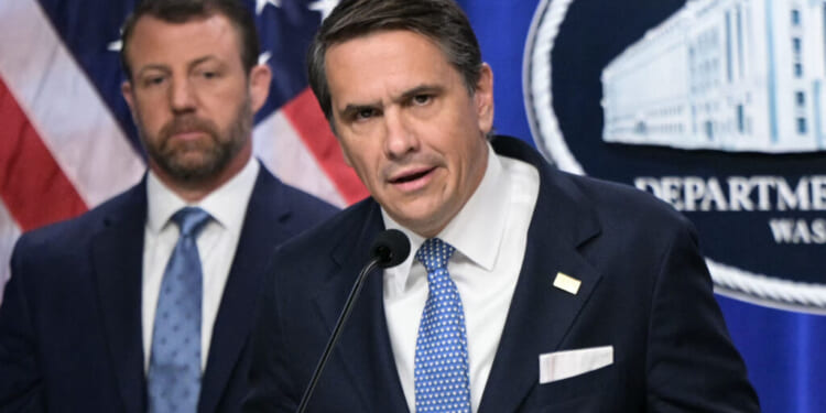 Todd Blanche and Markwayne Mullin in suits stand in front of U.S. flags and a sign that reads Department. The man in front speaks into a microphone, while the other looks on in the background.