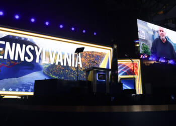 A stage with a large PENNSYLVANIA sign is lit up. A person stands in silhouette at the left. A screen shows another person speaking virtually, while empty seats are visible in the background.