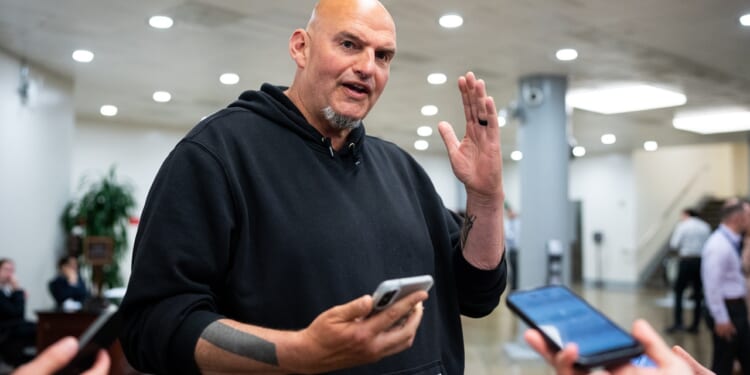 A bald man in a black hoodie gestures with one hand while holding a phone, surrounded by people holding smartphones in an indoor setting.