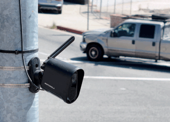 A black security camera is mounted on a metal pole beside a road, with a gray pickup truck driving by in the background.