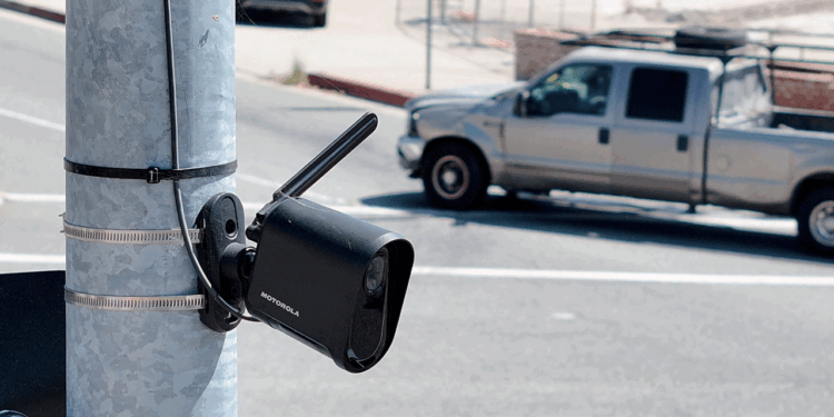 A black security camera is mounted on a metal pole beside a road, with a gray pickup truck driving by in the background.