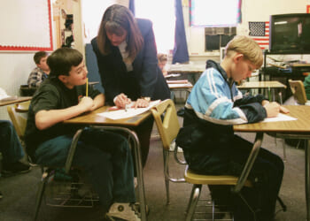 A teacher provides academic support to a student with his work in a classroom while other students sit at their desks, writing. An American flag and a bulletin board are visible in the background.