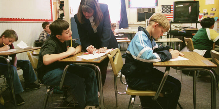 A teacher provides academic support to a student with his work in a classroom while other students sit at their desks, writing. An American flag and a bulletin board are visible in the background.
