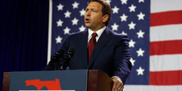 Ron DeSantis in a suit speaks at a podium labeled FREEDOM with a Florida outline, in front of a large American flag, emphasizing the importance of election integrity.