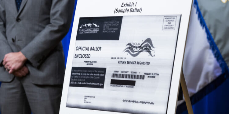 A sample ballot is displayed on an easel, labeled Exhibit 1, with a person in a suit standing nearby. The ballot includes official markings and return instructions.