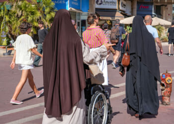 Women in hijabs walk outdoors, one guiding a person in a wheelchair. Other people in casual clothes walk nearby under bright sunlight in an urban setting with shops and palm trees visible.