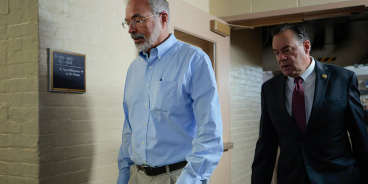 Two men walk through a hallway; one wears a light blue shirt and khaki pants, the other a dark suit and tie. The hallway has beige brick walls and a sign mounted on the left wall.
