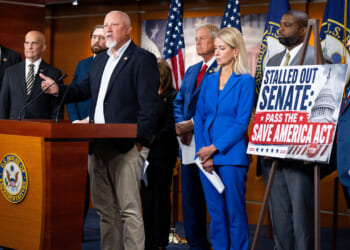 A group of people in business attire, including members of the Freedom Caucus, stand at a podium with U.S. flags. One man speaks while others listen intently. A sign reads, Stalled out Senate: Pass the Save America Act!.