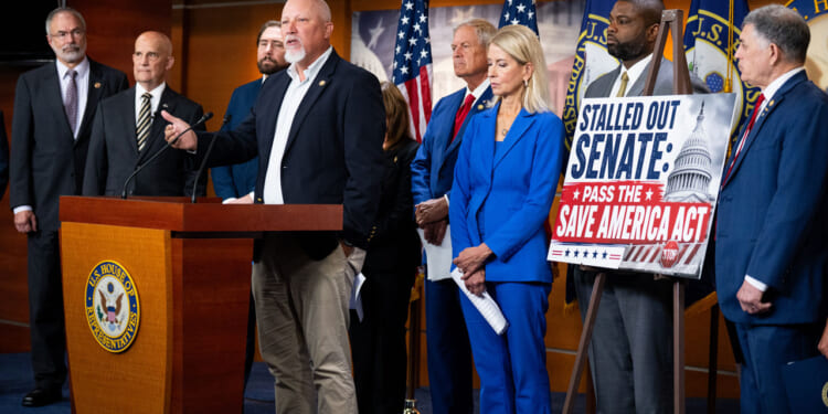 A group of people in business attire, including members of the Freedom Caucus, stand at a podium with U.S. flags. One man speaks while others listen intently. A sign reads, Stalled out Senate: Pass the Save America Act!.