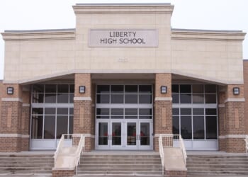 The front entrance of Liberty High School features wide steps, glass double doors, large windows, and a sign with the school name above the doors. The building exterior is made of tan and brown brick.