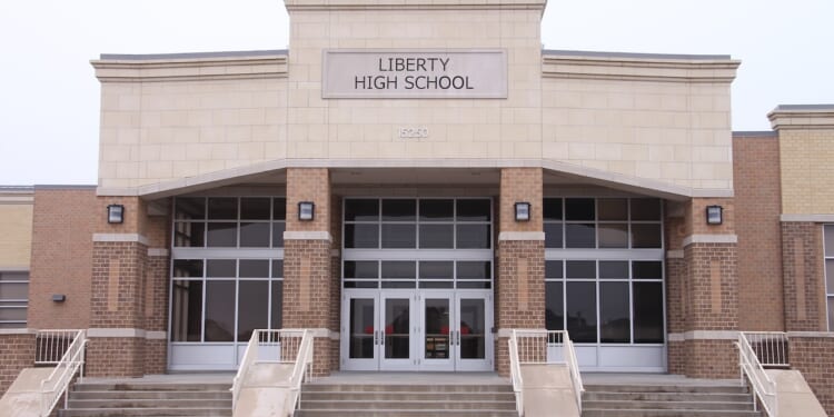 The front entrance of Liberty High School features wide steps, glass double doors, large windows, and a sign with the school name above the doors. The building exterior is made of tan and brown brick.