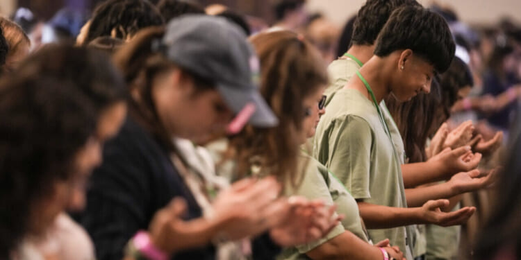 A group of people in casual clothing stand close together with heads bowed and hands open in front of them, appearing to be in prayer or deep reflection in a large indoor setting.