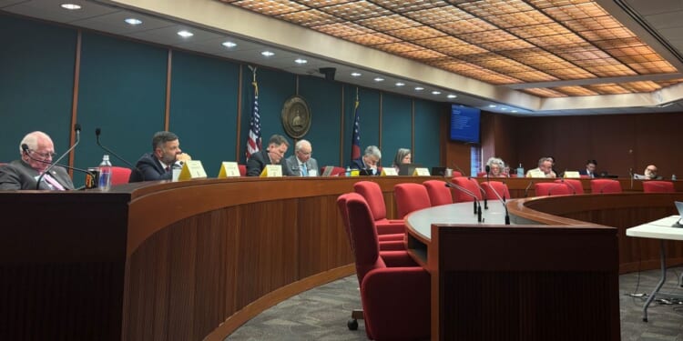 Members of the Joint Election Equipment Specifications and Standards Committee sit at a long, curved table at their third meeting with red chairs, U.S. and state flags, microphones, and nameplates.