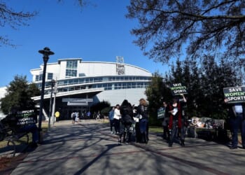 People holding Save Womens Sports and Support Women and Girls signs stand on a sidewalk outside a large, modern building on Georgia Tech's campus on a clear day; trees border the path.