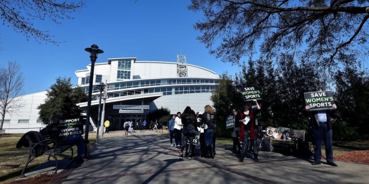 People holding Save Womens Sports and Support Women and Girls signs stand on a sidewalk outside a large, modern building on Georgia Tech's campus on a clear day; trees border the path.
