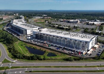 Aerial view of a large industrial data center under construction, surrounded by roads, parking lots, trees, and commercial buildings, with a clear blue sky overhead.