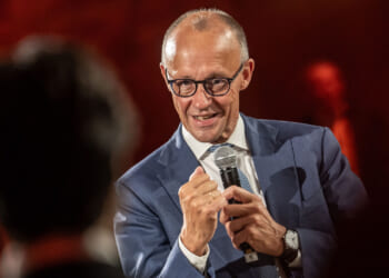 Friedrich Merz in a blue suit and glasses speaks into a microphone, gesturing with his fist raised, in front of a blurred, warmly lit background.