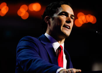 A man in a blue suit and red tie speaks at a podium against a dark background with orange lights blurred in the distance.
