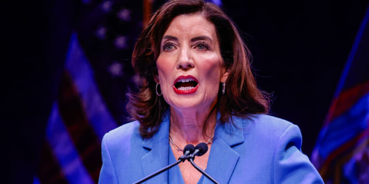 A woman in a blue blazer speaks at a podium with microphones, addressing the audience as she discusses the topic of pardon. She has shoulder-length brown hair and is shown against a dark background with blurred flags.