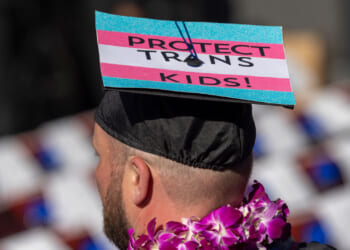 A person wearing a graduation cap decorated with the trans pride colors and the words Protect Trans Kids! stands among a crowd, with a purple flower lei around their neck. Teachers are being taught to indoctrinate according to a new study.
