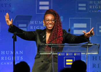 Human Rights Campaign President Kelley Robinson, a person with long red braids and glasses smiles and gestures with open arms while standing at a podium, against a blue Human Rights Campaign backdrop.