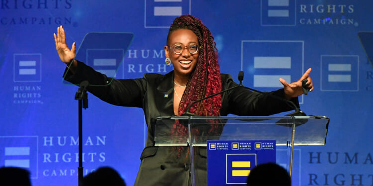 Human Rights Campaign President Kelley Robinson, a person with long red braids and glasses smiles and gestures with open arms while standing at a podium, against a blue Human Rights Campaign backdrop.