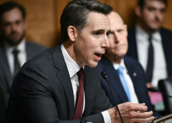 A man in a suit speaks into a microphone at a formal hearing, with other people seated and listening in the background.