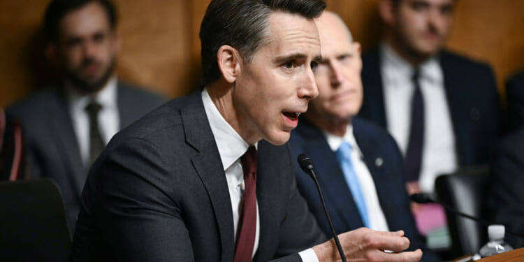 A man in a suit speaks into a microphone at a formal hearing, with other people seated and listening in the background.