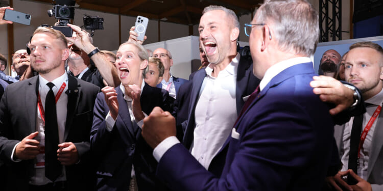 A group of people from the AfD in business attire cheer and celebrate together indoors. Some are smiling and shouting, while others take photos. The atmosphere is lively and joyful.
