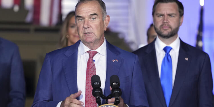 A man in a blue suit and red tie speaks at a podium with microphones, while another man in a blue suit and blue tie stands behind him. American flags are visible in the background.