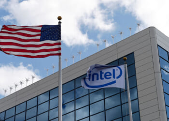 An American flag and an Intel flag wave in front of a modern glass office building with a cloudy sky in the background.