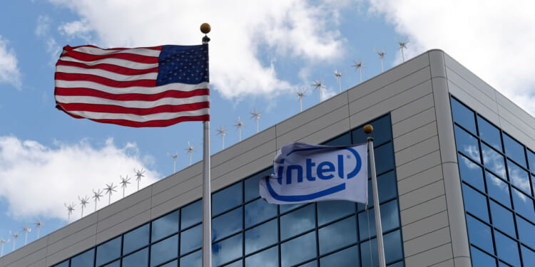An American flag and an Intel flag wave in front of a modern glass office building with a cloudy sky in the background.