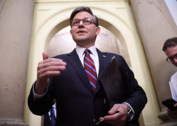 A man in a suit speaks, holding a folder, while two others stand nearby using their phones in a hallway with large columns and an arched doorway.
