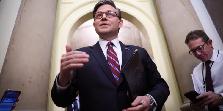 A man in a suit speaks, holding a folder, while two others stand nearby using their phones in a hallway with large columns and an arched doorway.