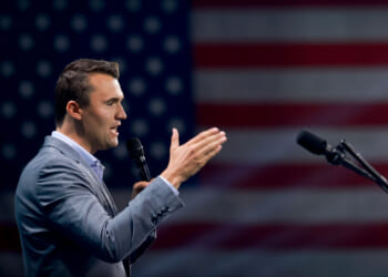 Charlie Kirk in a gray suit speaks into a microphone with an American flag in the blurred background.