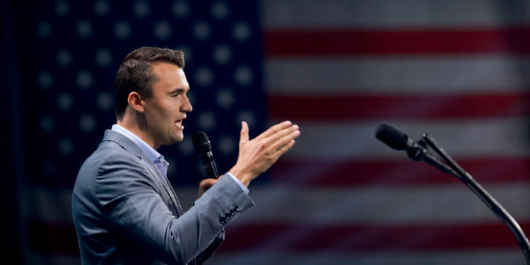 Charlie Kirk in a gray suit speaks into a microphone with an American flag in the blurred background.
