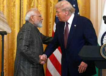 Donald Trump and Indian Prime Minister Narendra Modi in suits shake hands at a podium with the U.S. and Indian flags behind them. The setting appears formal, likely a press conference or official meeting.