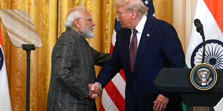 Donald Trump and Indian Prime Minister Narendra Modi in suits shake hands at a podium with the U.S. and Indian flags behind them. The setting appears formal, likely a press conference or official meeting.