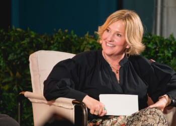 A woman with short blonde hair sits in a beige chair, smiling and holding a card. She wears a black blouse, floral skirt, and a headset microphone. Greenery and blurred background elements are visible behind her.